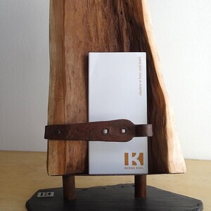 Peut inclure: Un porte-cartes en bois avec un fini grain de bois naturel. Il comporte une bande métallique rouillée et deux pieds en métal, reposant sur une base en ardoise noire. Le support affiche une carte blanche avec le texte "BERND KOHL".