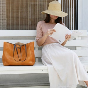 May include: A woman wearing a straw hat and a white skirt sits on a white bench holding a book and a brown leather tote bag with a monogram.