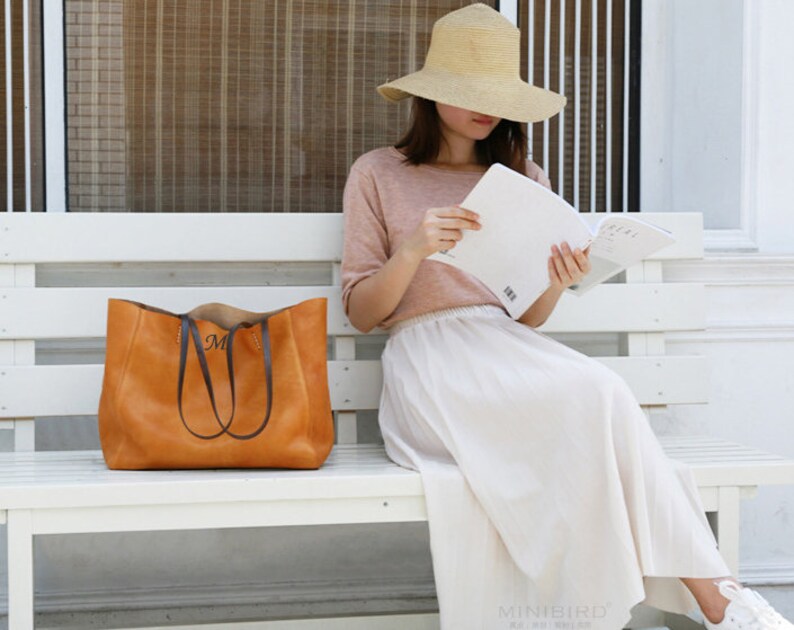 May include: A woman wearing a straw hat and a white skirt sits on a white bench holding a book and a brown leather tote bag with a monogram.