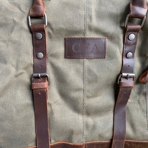 May include: An olive green canvas duffel bag featuring dark brown leather straps with metal buckles and rivets. A rectangular leather patch is centered on the front with the embossed letters CFA. A vertical zippered pocket is located on the right side.