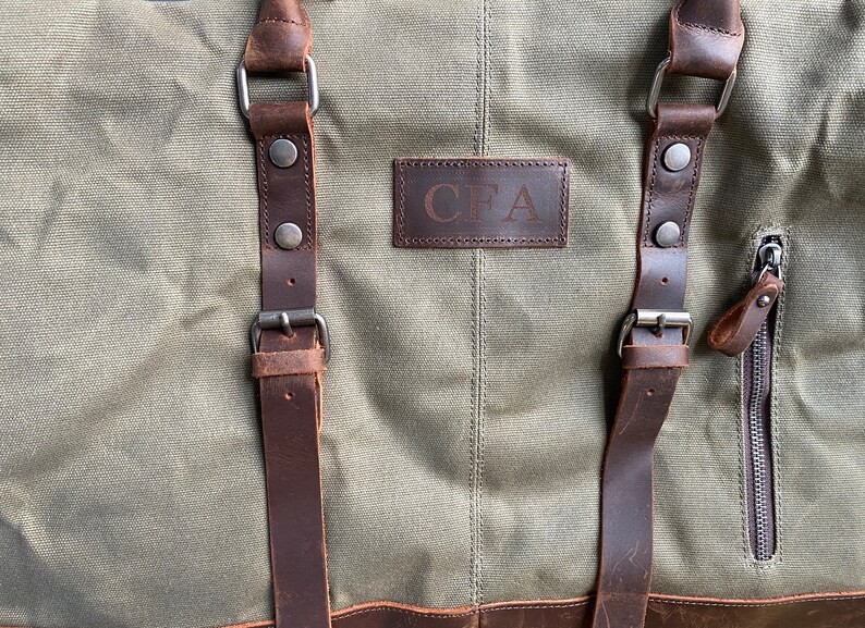 May include: An olive green canvas duffel bag featuring dark brown leather straps with metal buckles and rivets. A rectangular leather patch is centered on the front with the embossed letters CFA. A vertical zippered pocket is located on the right side.