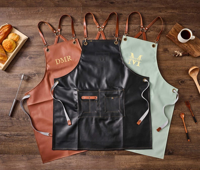 May include: Three leather aprons with adjustable straps. The aprons are brown, black, and light green. The brown apron has the initials "DMR" embroidered on the front. The black apron has a small square with the initials "DM" embroidered on the front. The light green apron has the initial "M" embroidered on the front.