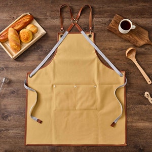 May include: A tan canvas apron with brown leather straps and a large pocket. The apron has a criss-cross strap design at the back.