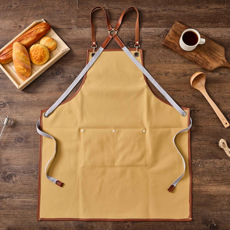 May include: A tan canvas apron with brown leather straps and a large pocket. The apron has a criss-cross strap design at the back.