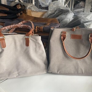 May include: Two rectangular handbags made of light gray canvas material. Each bag features two brown faux leather handles attached with gold-toned metal hardware. The left bag has a protective plastic wrap on the handles, while the right bag has a rectangular brown faux leather patch on the front.