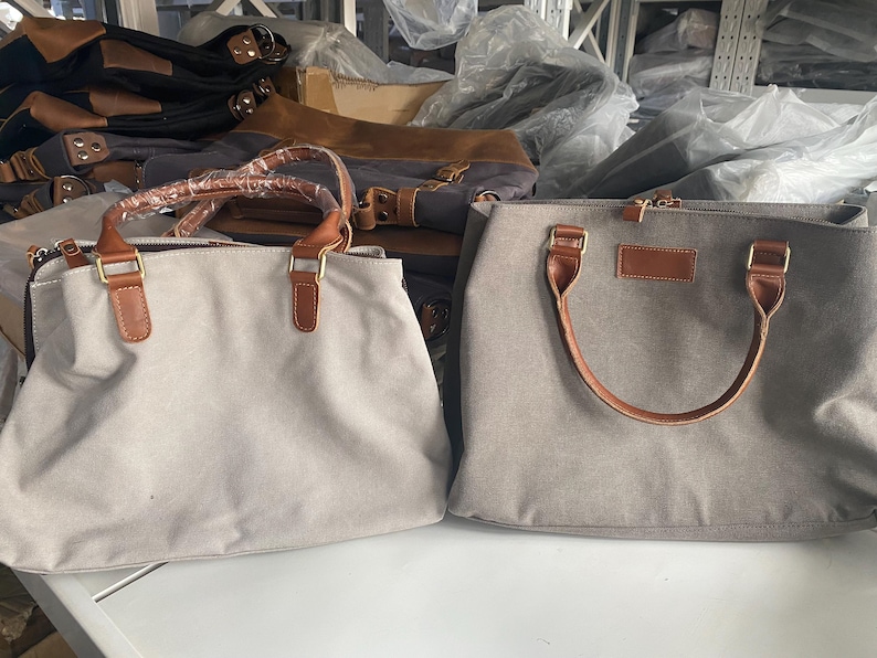 May include: Two rectangular handbags made of light gray canvas material. Each bag features two brown faux leather handles attached with gold-toned metal hardware. The left bag has a protective plastic wrap on the handles, while the right bag has a rectangular brown faux leather patch on the front.