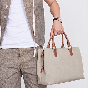 May include: A beige canvas handbag featuring brown leather handles and trim, a top zipper closure with leather pulls, and metal hardware. The bag is rectangular in shape with a structured base and is shown alongside a white measuring tape.