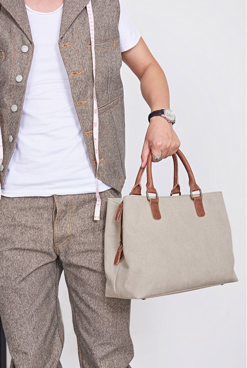 May include: A beige canvas handbag featuring brown leather handles and trim, a top zipper closure with leather pulls, and metal hardware. The bag is rectangular in shape with a structured base and is shown alongside a white measuring tape.
