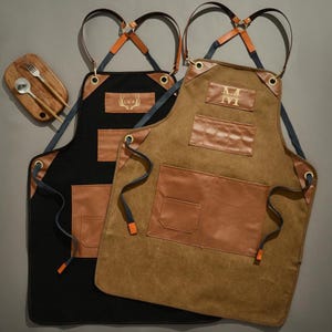 Op de afbeelding: Twee schorten, één zwart en één bruin, met leren accenten en zakken. De schorten hebben verstelbare bandjes en metalen oogjes. Een houten serveerplank met vork en lepel staat linksboven. Op het bruine schort staat de letter "M".