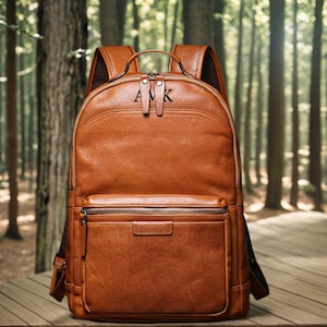 Op de afbeelding: Een bruine leren rugzak met een voorvak met ritssluiting en een handvat aan de bovenkant. De rugzak heeft de initialen "AMK" in reliëf op de bovenkant. De achtergrond is een vage bosscène.