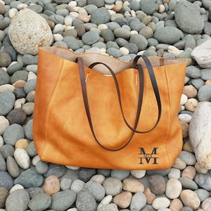 May include: A large, brown leather tote bag with a brown leather strap. The bag has a monogram on the front that reads "MIA M".