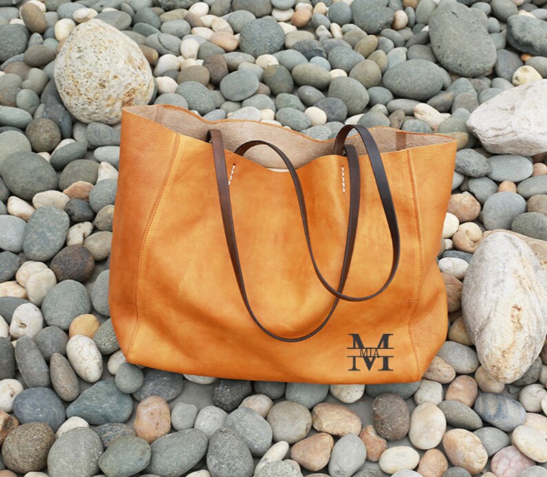 May include: A large, brown leather tote bag with a brown leather strap. The bag has a monogram on the front that reads "MIA M".
