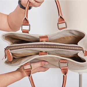 May include: A beige canvas tote bag featuring brown leather handles attached with brass-toned square hardware. The bag has an open top design with a central zippered compartment, brown interior lining, and a small leather tab closure with a snap.