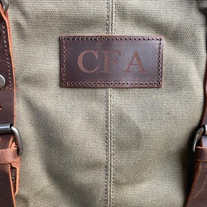 May include: An olive green canvas bag featuring dark brown leather straps with silver-toned metal buckles and rivets. A rectangular dark brown leather patch is stitched onto the center, embossed with the capital letters CFA in a serif font.