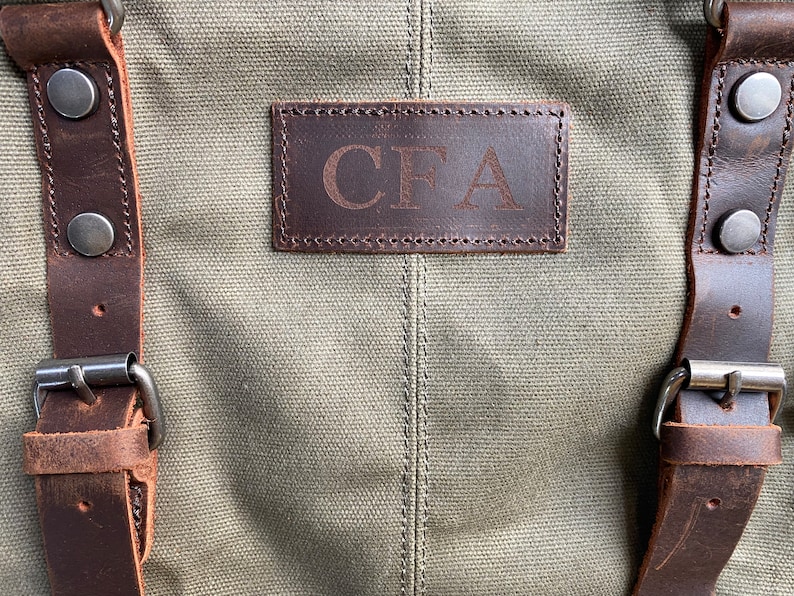 May include: An olive green canvas bag featuring dark brown leather straps with silver-toned metal buckles and rivets. A rectangular dark brown leather patch is stitched onto the center, embossed with the capital letters CFA in a serif font.