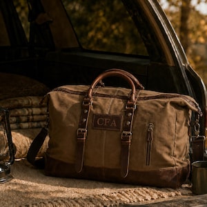 May include: A brown canvas duffel bag with dark brown leather trim, two top handles, and adjustable leather straps with metal buckles. A rectangular leather patch on the front is embossed with the letters CFA. It features a side zipper pocket.