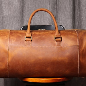 May include: A brown leather duffel bag with two top handles and a zipper closure. The bag is sitting on a wooden stool.