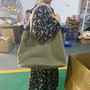 May include: A large olive green canvas tote bag with brown leather-like shoulder straps. The bag features a wide, rectangular body with a flat base and reinforced stitching at the strap attachments. The shoulder strap is partially covered in plastic.