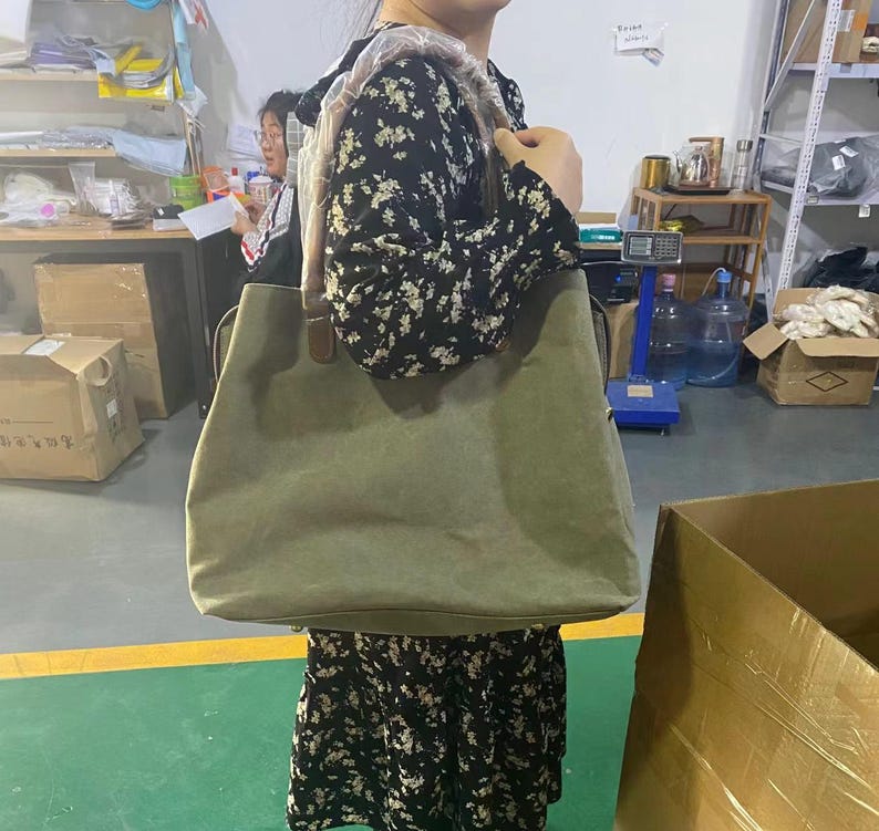 May include: A large olive green canvas tote bag with brown leather-like shoulder straps. The bag features a wide, rectangular body with a flat base and reinforced stitching at the strap attachments. The shoulder strap is partially covered in plastic.
