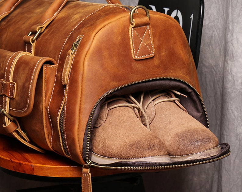 May include: A brown leather duffel bag with a zippered opening. The bag is open and two pairs of brown suede shoes are visible inside.