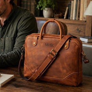 May include: A tan leather briefcase featuring two top handles, a detachable shoulder strap with a metal buckle, and the letters MHK embossed on the front. The bag includes brass-colored hardware, reinforced stitching, and a structured rectangular shape.