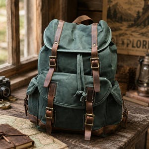 May include: A dark green canvas backpack featuring brown leather straps with brass-toned metal buckles. The bag includes a large main compartment with a flap closure, a front pocket with a flap, and two side pockets, all secured with leather straps.