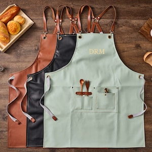 May include: Three aprons with adjustable straps. The apron on the right is light green with two pockets and the text "DRM" embroidered on the front. The apron in the middle is black leather and the apron on the left is brown leather.