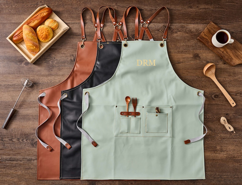 May include: Three aprons with adjustable straps. The apron on the right is light green with two pockets and the text "DRM" embroidered on the front. The apron in the middle is black leather and the apron on the left is brown leather.