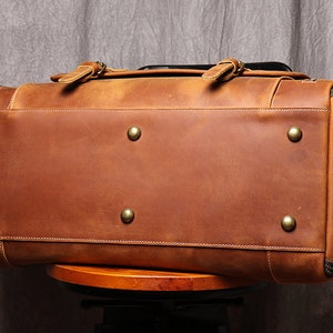 May include: A brown leather duffel bag with brass accents. The bag is sitting on a wooden stool.