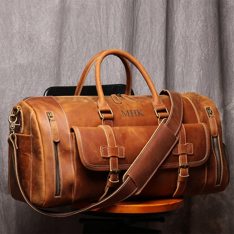May include: A brown leather duffel bag with a long shoulder strap and a top handle. The bag has a front pocket and the initials "MHK" are embossed on the front.
