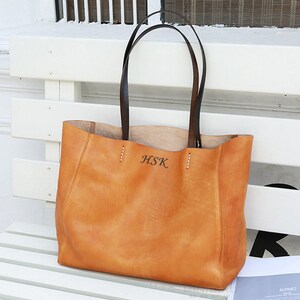 May include: A tan leather tote bag with black leather handles. The bag has a stitched design and the initials "HSK" are embossed on the front.