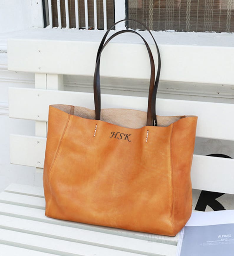 May include: A tan leather tote bag with black leather handles. The bag has a stitched design and the initials "HSK" are embossed on the front.