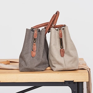 May include: Two canvas handbags, one in dark gray and one in light beige, featuring brown leather handles and trim. Each bag has a vertical zippered side pocket with a brown leather pull tab and a metallic zipper closure, showcasing a structured design.