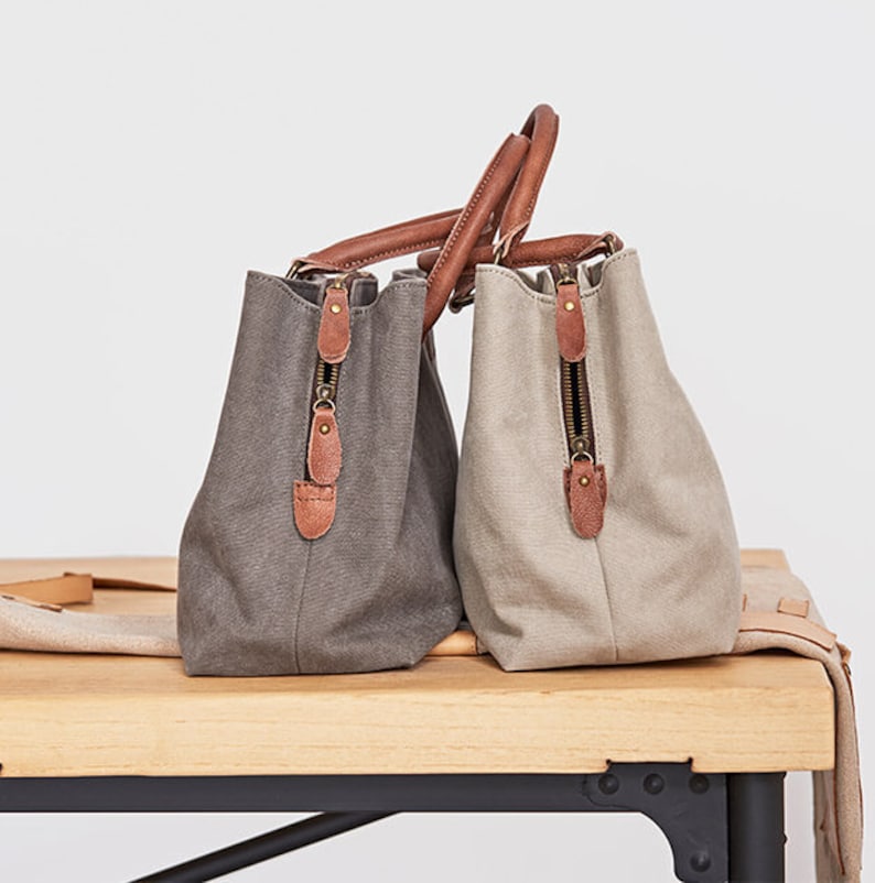 May include: Two canvas handbags, one in dark gray and one in light beige, featuring brown leather handles and trim. Each bag has a vertical zippered side pocket with a brown leather pull tab and a metallic zipper closure, showcasing a structured design.