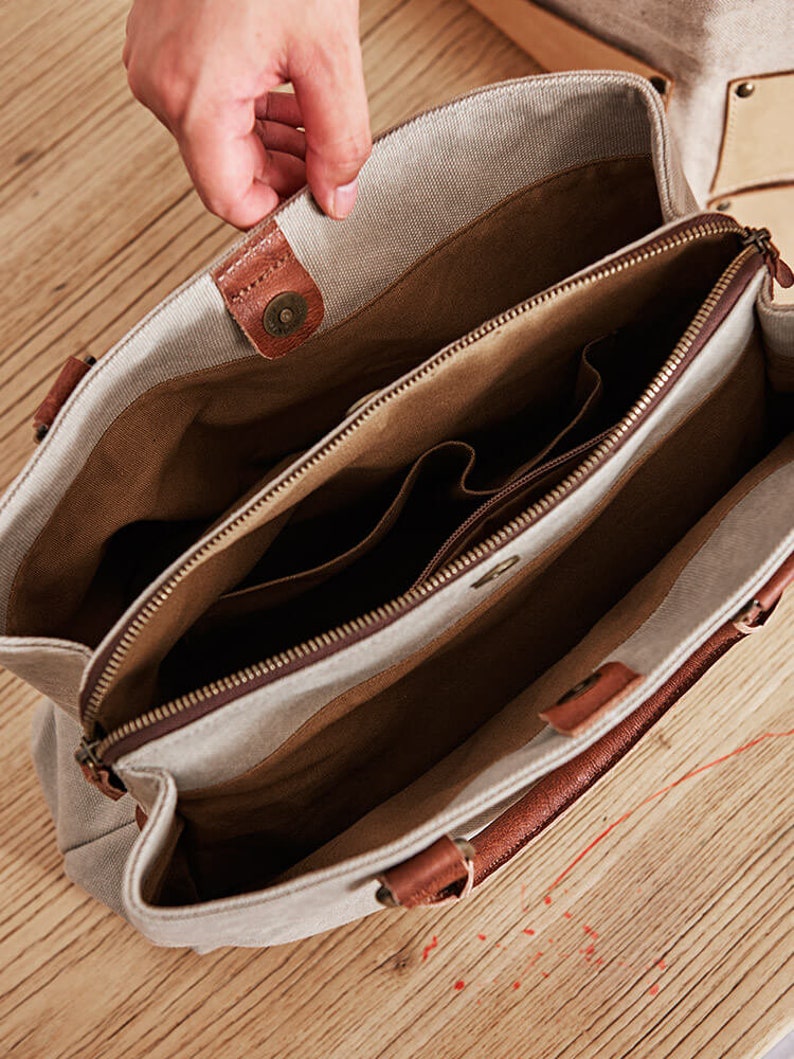 May include: A beige canvas tote bag with brown leather accents and handles. The interior is lined with brown fabric and features a central zippered compartment, multiple open pockets, and a magnetic snap closure at the top opening of the bag.