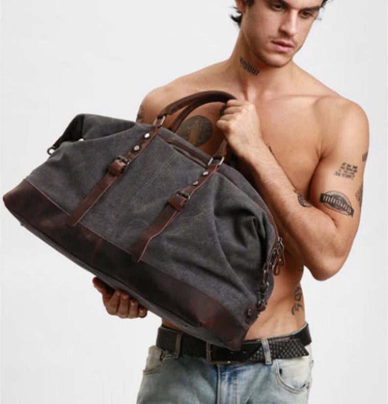 May include: A charcoal gray canvas duffel bag featuring dark brown leather trim, two vertical leather straps with metal buckles, a top handle, and metal hardware accents. The bag has a structured base and a spacious main compartment for storage.