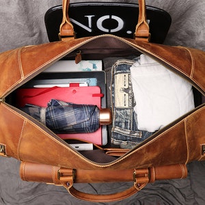May include: A brown leather duffel bag with a zippered opening. The bag is open and filled with various items, including a plaid umbrella, a red folder, a white t-shirt, and blue jeans.
