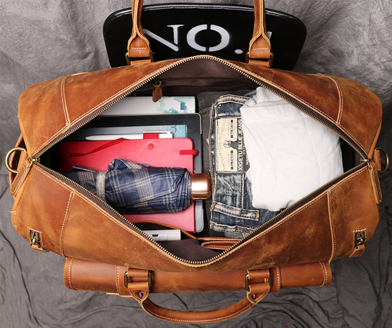 May include: A brown leather duffel bag with a zippered opening. The bag is open and filled with various items, including a plaid umbrella, a red folder, a white t-shirt, and blue jeans.