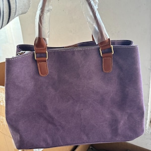 May include: A purple canvas handbag featuring brown faux leather handles and gold-toned metal hardware. The handles are wrapped in protective clear plastic. The bag has a rectangular shape with visible stitching along the top edge and a zipper closure.