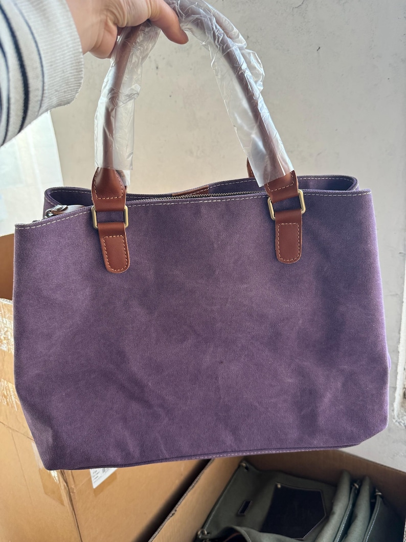 May include: A purple canvas handbag featuring brown faux leather handles and gold-toned metal hardware. The handles are wrapped in protective clear plastic. The bag has a rectangular shape with visible stitching along the top edge and a zipper closure.