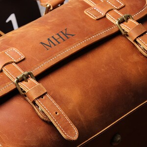May include: A brown leather duffel bag with a monogrammed inscription "MHK" on the front. The bag has two adjustable straps with brass buckles.