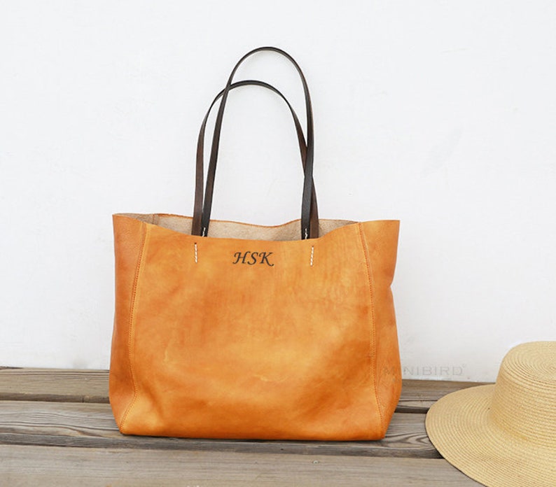 May include: A tan leather tote bag with brown leather handles. The bag has a monogrammed "HSK" on the front.