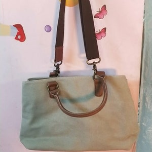 May include: A light sage green canvas handbag featuring brown faux leather handles and a matching adjustable brown fabric shoulder strap. The bag includes metallic hardware clips for the strap and a top zipper closure for the main compartment.