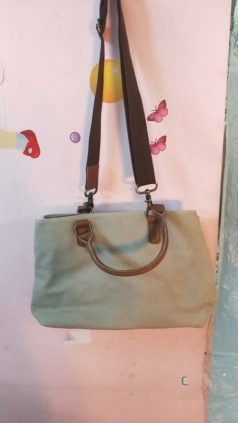 May include: A light sage green canvas handbag featuring brown faux leather handles and a matching adjustable brown fabric shoulder strap. The bag includes metallic hardware clips for the strap and a top zipper closure for the main compartment.