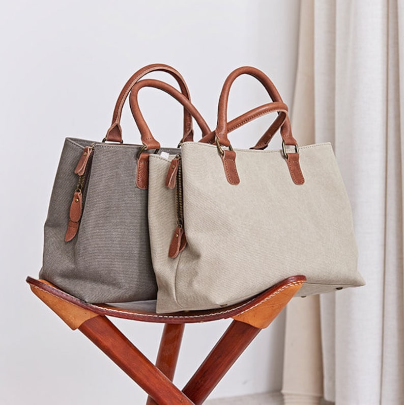 May include: Two structured handbags made of textured canvas fabric, one in dark gray and one in light beige. Each bag features two brown leather handles, a side zipper closure with a leather pull tab, and metal hardware connecting the handles.