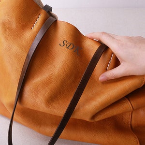 May include: A large, brown leather tote bag with two long straps. The bag has a subtle, textured finish and the initials "SDK" are embossed on the front.