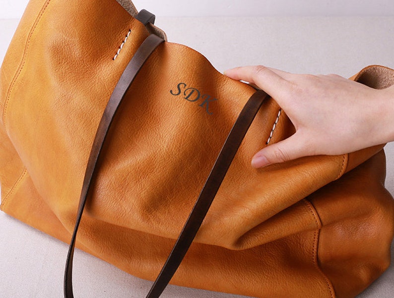 May include: A large, brown leather tote bag with two long straps. The bag has a subtle, textured finish and the initials "SDK" are embossed on the front.