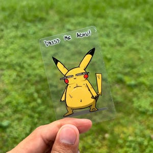 Könnte beinhalten: Ein transparenter Aufkleber mit einer gelben Cartoonfigur mit Sonnenbrille und schwarzen Ohrenspitzen. Der Text "DRESS ME ACHU!" steht über der Figur. Die Figur hat einen mürrischen Gesichtsausdruck.