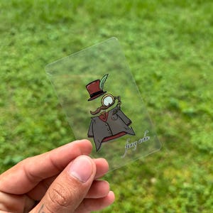 Könnte beinhalten: Ein klarer, rechteckiger Aufkleber mit einer Cartoon-Illustration eines Herrn mit Zylinder, Monokel und Schnurrbart. Der Herr trägt eine graue Jacke und eine rote Weste. Der Aufkleber hat den Text "fancy achu" in Kursivschrift.