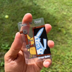 Könnte beinhalten: Eine transparente rechteckige Karte mit einem Cartoon-Pikachu-Design und dem Text "Dress Me Achu!" und "X-ray Achu." Die Karte zeigt Pikachu in einer Röntgenansicht, mit einem Kaninchenskelett. Die Karte wird in einer Hand vor grünem Hintergrund gehalten.